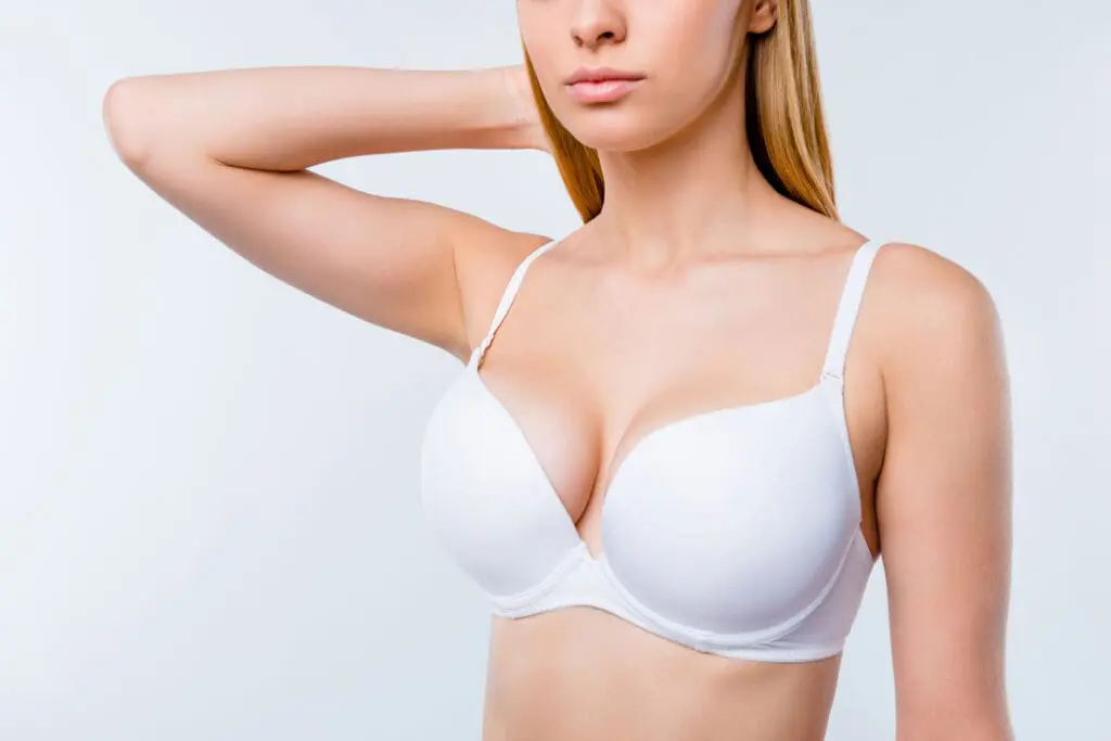 Cropped close-up portrait of nice calm confident attractive lovely blonde girl chest after uplift injection wearing bra touching hair isolated over light gray background Breast Augmentation in West Palm Beach and Jupiter, FL