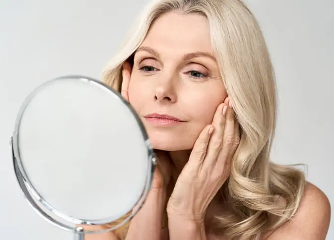 Closeup portrait of gorgeous happy middle age woman looking at mirror touching her skin enjoying treatment for dry skin. Advertising of antiaging beauty skin care products. Silhouette Instalift in West Palm Beach and Jupiter, FL